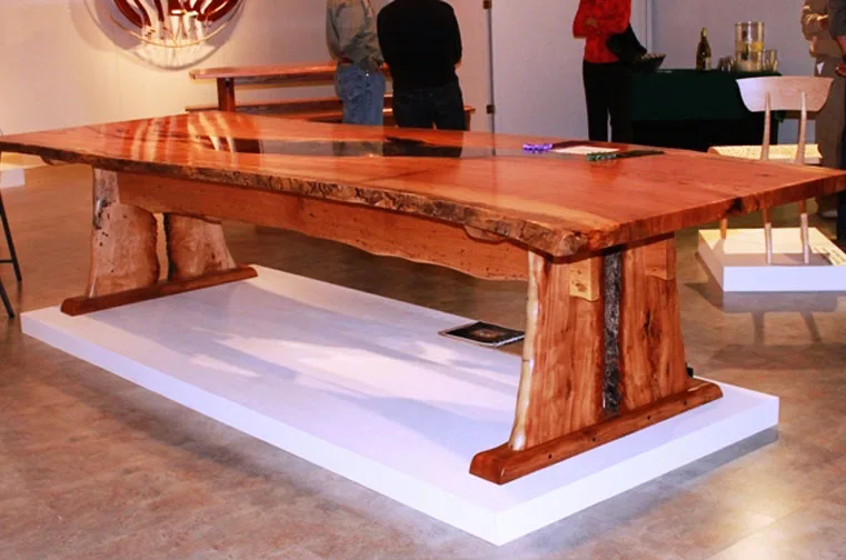 Live-edge wood slab dining table with natural bark edges and sculptural pedestal base displayed on white platform in modern showroom setting.