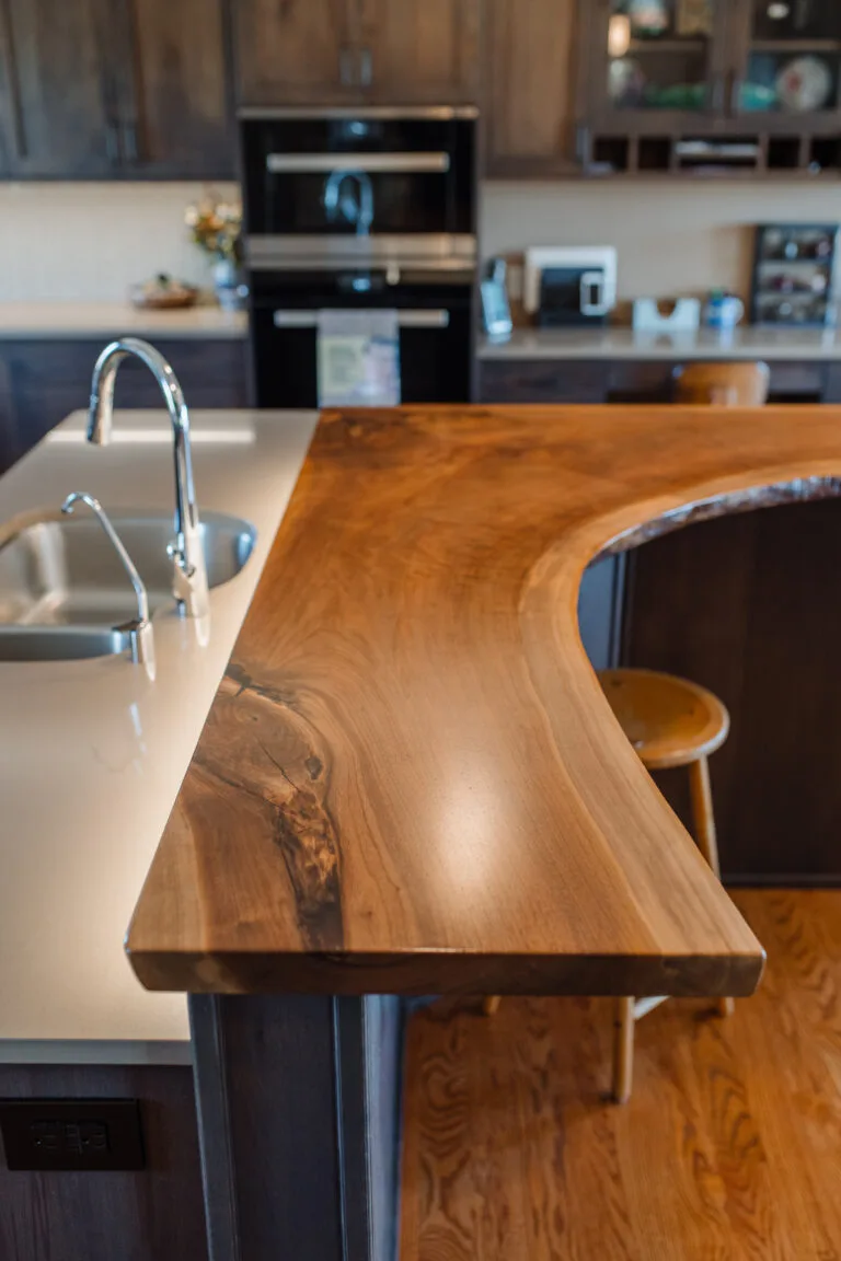 Live-edge wood slab countertop with natural curves and knot details extends from modern kitchen island with integrated sink and seating area.