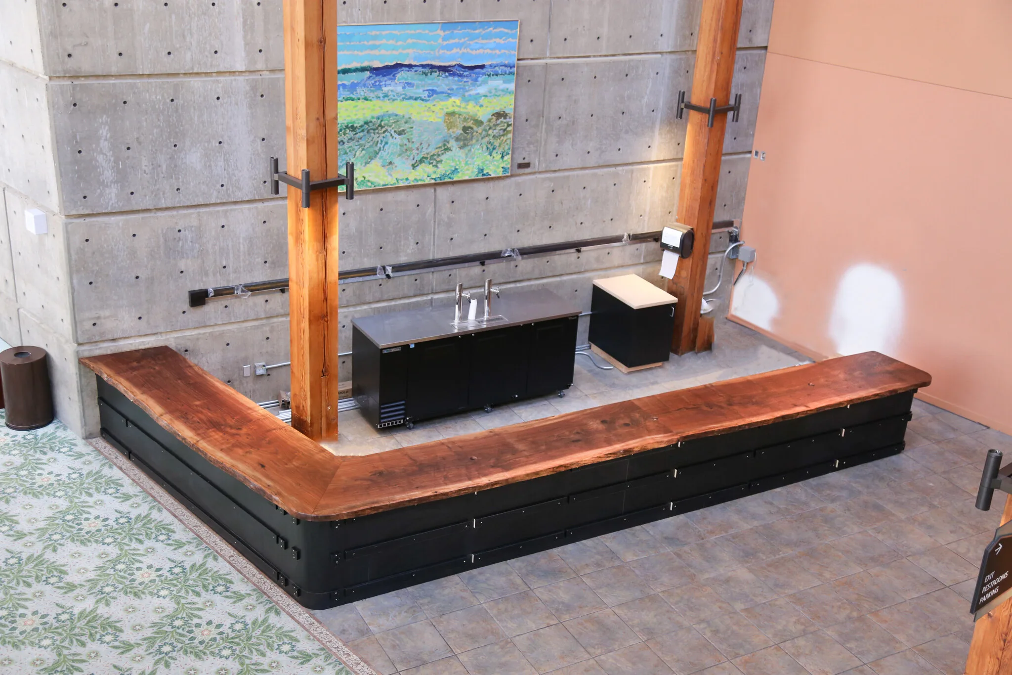 Large L-shaped live-edge wood slab bar top with natural edge detail mounted on black metal base in modern showroom with concrete walls and exposed beams.