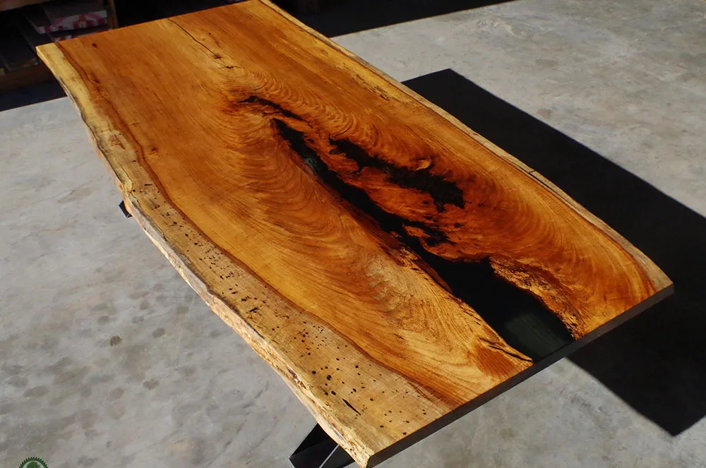 Live-edge wood slab with dramatic natural void and rich grain patterns displayed on black metal base in workshop setting.