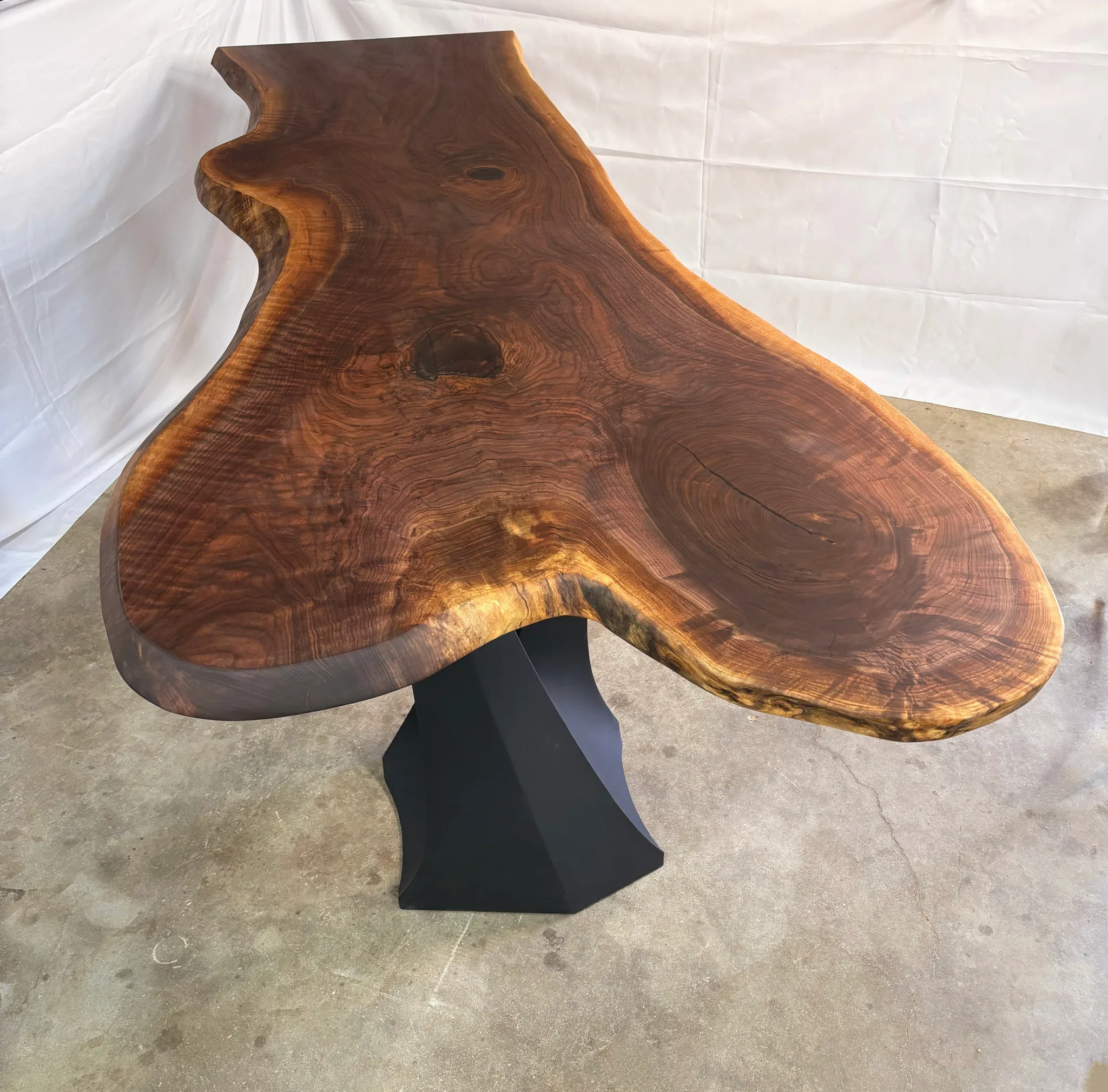 Dramatic live-edge walnut slab table with organic flowing shape and rich grain patterns mounted on modern geometric black metal base.