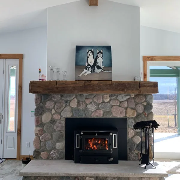 Rustic reclaimed wood mantel beam anchors a stone fireplace surround in a bright, vaulted living room with natural light and modern farmhouse details.