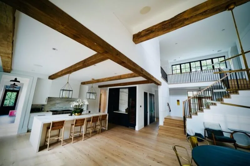 Dramatic exposed wood beams span a vaulted ceiling above light wood flooring in an open-concept kitchen and living space with modern farmhouse design.