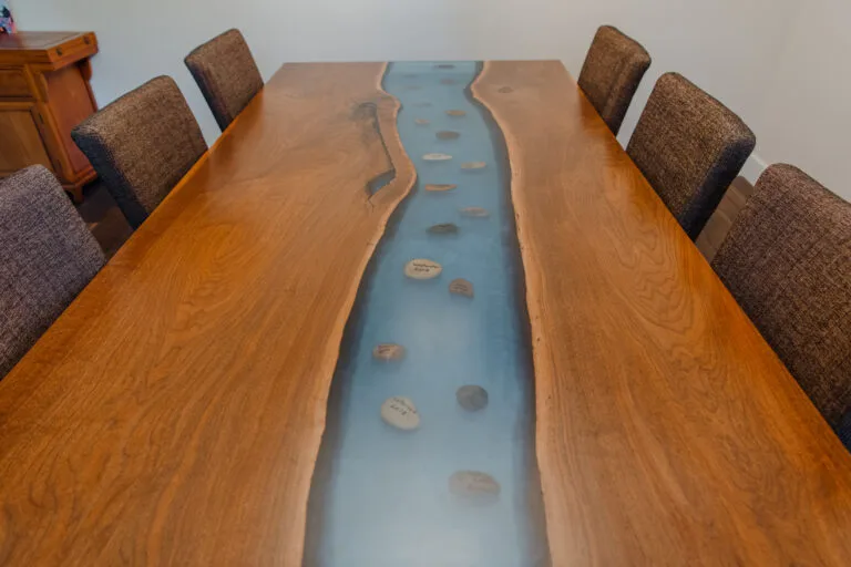 Live-edge wood dining table featuring a striking blue epoxy river inlay with embedded stones running through the center of two natural-edge slabs.