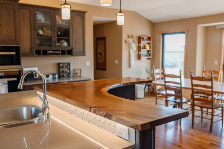 Live-edge wood slab countertop wraps around kitchen island with integrated cooktop, showcasing natural edge and warm grain against modern cabinetry.