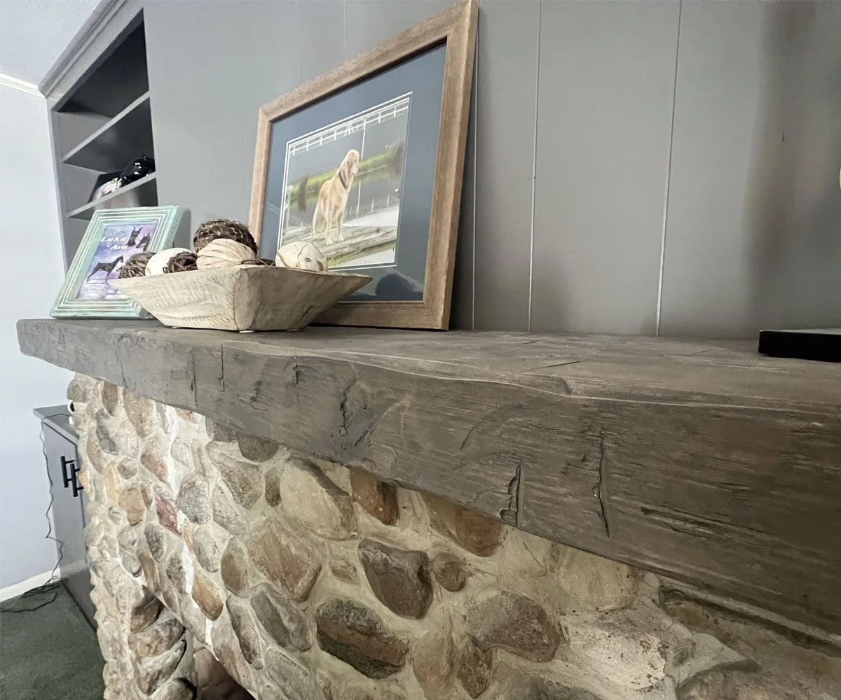 Weathered reclaimed wood mantel with visible mortise joints mounted on stone fireplace surround, styled with rustic decor and framed artwork.