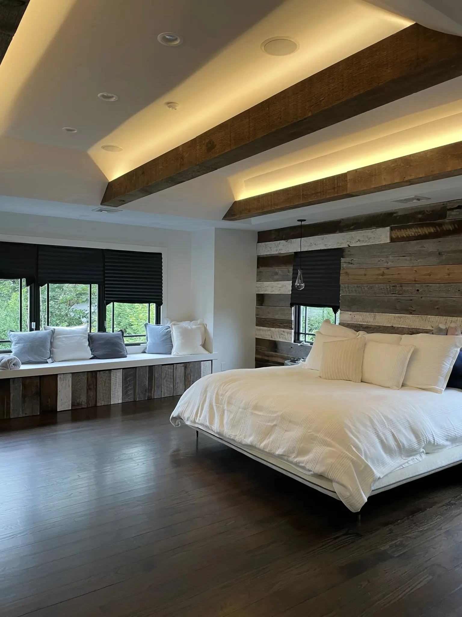 Modern bedroom featuring dramatic exposed ceiling beams with uplighting, reclaimed wood accent wall, and dark hardwood flooring creating a sophisticated rustic aesthetic.