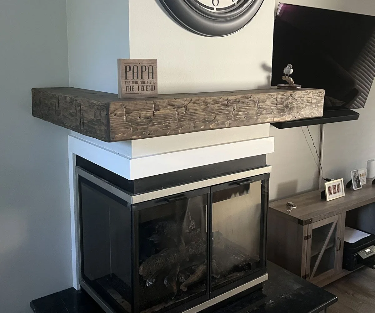 Rustic reclaimed wood fireplace mantel with distressed gray finish and hand-hewn texture mounted above modern glass-front fireplace insert.