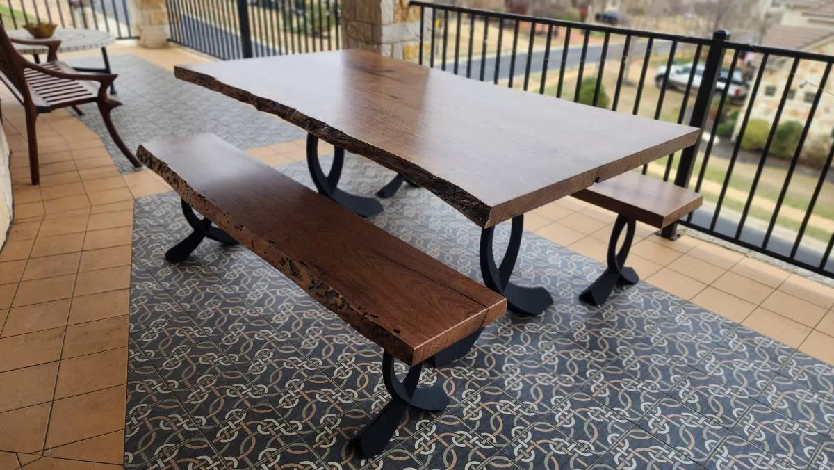 Live-edge wood slab dining table with matching bench and side table on covered outdoor patio, featuring black curved metal legs.