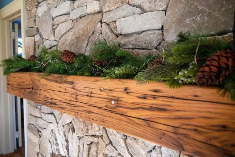 Rustic reclaimed wood mantel with distressed finish and nail holes mounted on stacked stone fireplace, decorated with evergreen garland and pinecones.