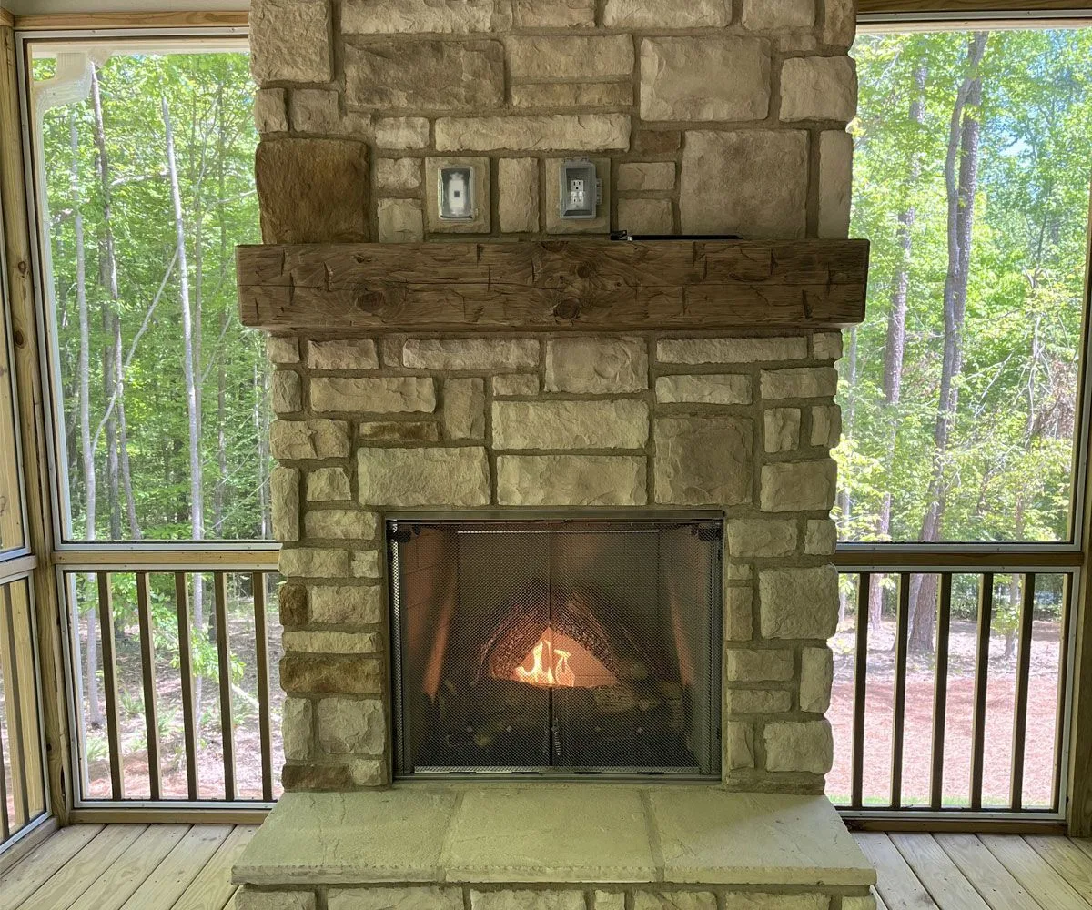 Rustic reclaimed wood mantel anchors a stone fireplace in a screened porch overlooking wooded landscape, featuring natural grain and hand-hewn texture.